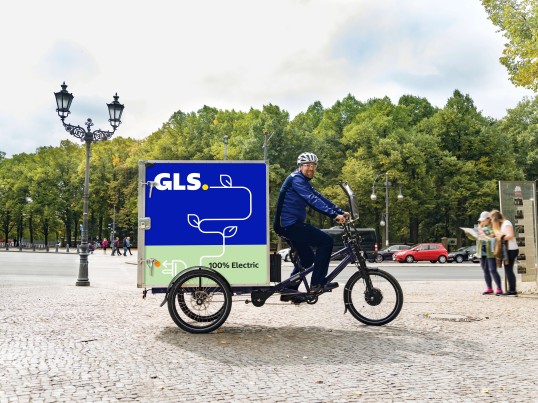 GLS futár elektromos teherkerékpárral áthajt egy parkon.